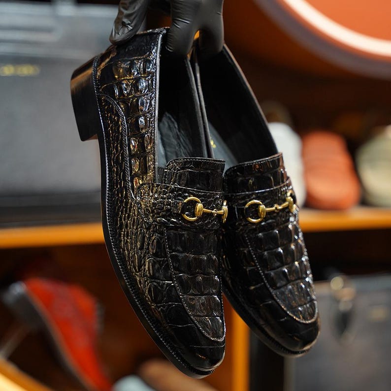 May include: A pair of black leather loafers with a crocodile skin texture. The shoes have a gold-colored metal bit detail on the vamp. The shoes are held up by a gloved hand.