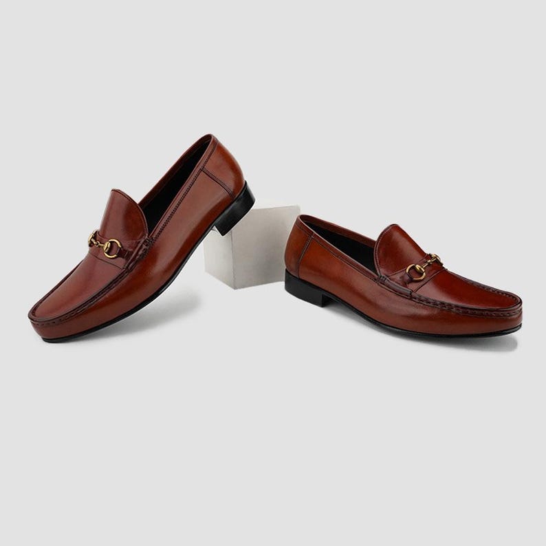 May include: A pair of brown leather loafers with a gold-colored metal accent on the vamp. The shoes have a classic slip-on design with a square toe and a black sole. The loafers are displayed on a white surface.