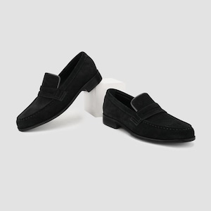 May include: Two black suede leather loafers. The shoes have a classic slip-on design with a low heel and a strap across the top. The shoes are displayed on a white surface against a neutral background.