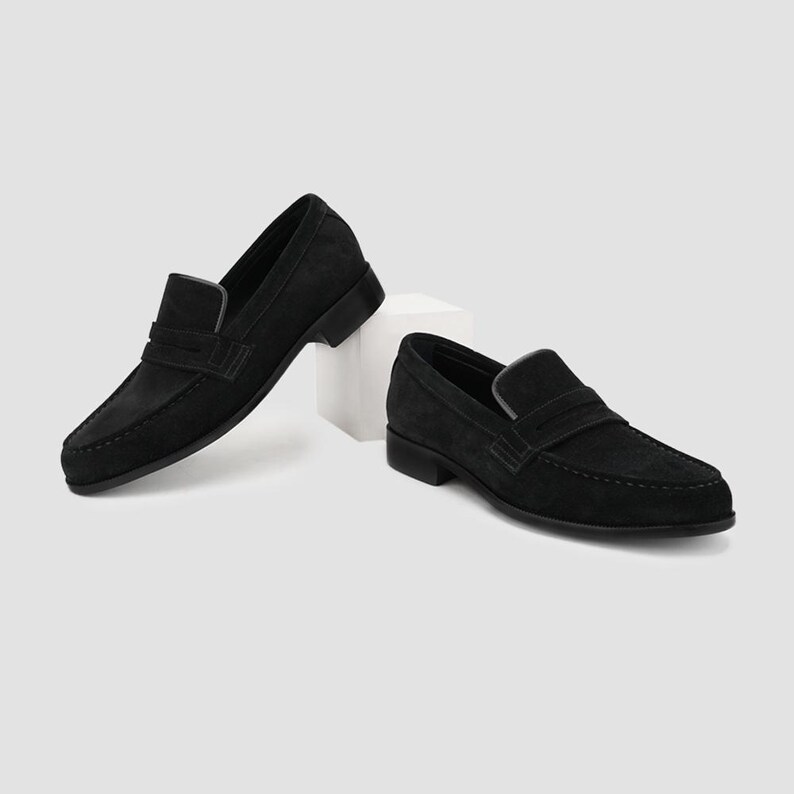May include: Two black suede leather loafers. The shoes have a classic slip-on design with a low heel and a strap across the top. The shoes are displayed on a white surface against a neutral background.