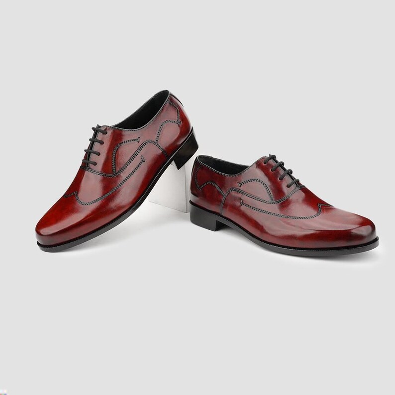 May include: A pair of burgundy leather dress shoes with black laces. The shoes have a wingtip design and a glossy finish. The shoes are displayed on a white surface against a light gray background.