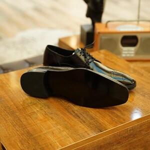 Puede incluir: Un par de zapatos de vestir de cuero negro con un acabado brillante, exhibidos sobre una superficie de madera. Los zapatos tienen un dise&ntilde;o cl&aacute;sico con cordones y est&aacute;n colocados cerca de una radio de estilo vintage.
