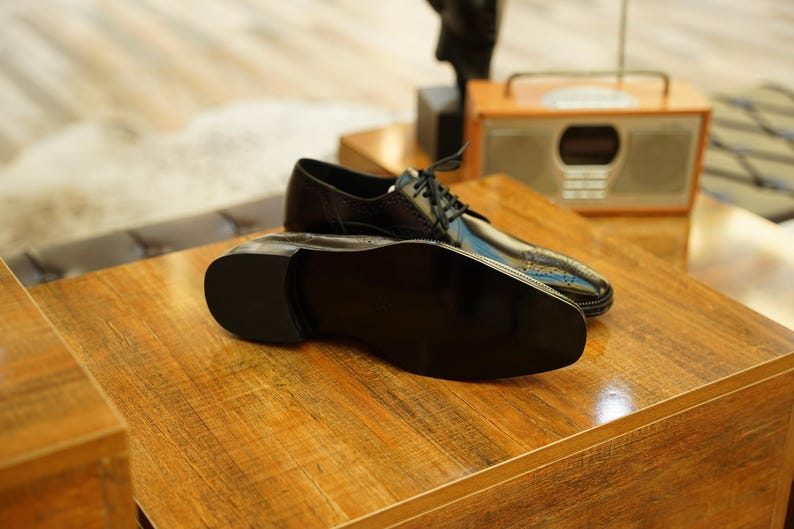 Puede incluir: Un par de zapatos de vestir de cuero negro con un acabado brillante, exhibidos sobre una superficie de madera. Los zapatos tienen un dise&ntilde;o cl&aacute;sico con cordones y est&aacute;n colocados cerca de una radio de estilo vintage.