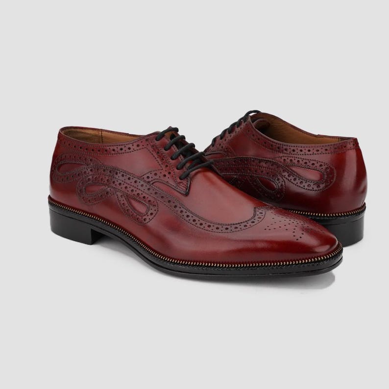 Puede incluir: Zapatos de vestir de cuero burdeos con cordones negros. Los zapatos presentan perforaciones decorativas y un dise&ntilde;o detallado en los laterales. Las suelas son negras con un fino ribete dorado.