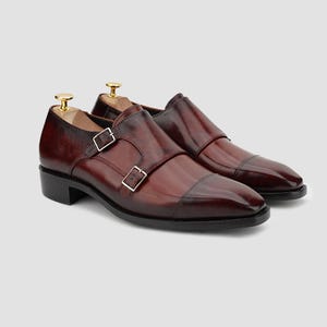 Può includere: Un paio di scarpe doppie monk in pelle bordeaux. Le scarpe presentano un design a punta, fibbie argentate e un tacco basso. Sono presenti le forme per le scarpe. La pelle è lucida.