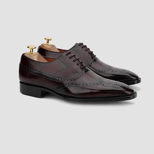 Può includere: Un paio di scarpe eleganti in pelle bordeaux con dettagli brogue. Le scarpe presentano un classico design a punta di ala, lacci marrone scuro e una finitura lucida. Le scarpe sono esposte con forme di legno. Le scarpe misurano circa 30 cm.