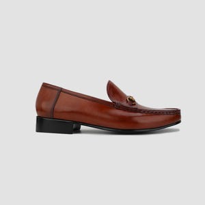 May include: A brown leather loafer with a gold-colored metal accent. The shoe has a low black heel and a classic design. The leather has a glossy finish.