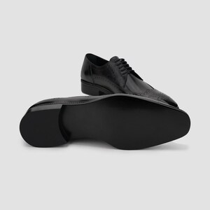 Puede incluir: Zapatos de vestir de cuero negro con detalles brogue y acabado brillante. Un zapato est&aacute; en posici&oacute;n vertical, mientras que el otro muestra la suela. Dise&ntilde;o cl&aacute;sico y formal.