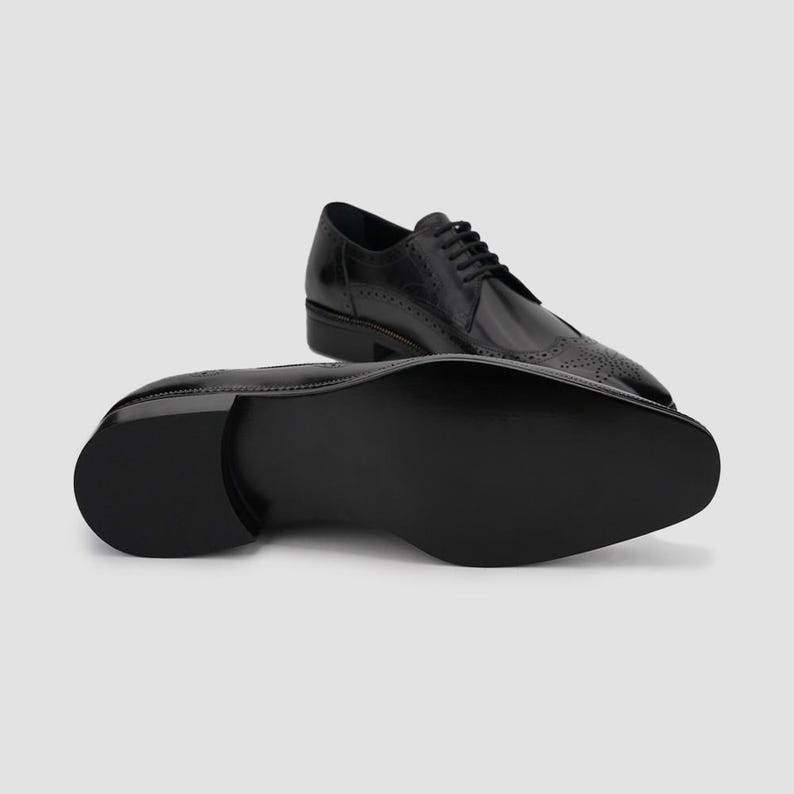 Puede incluir: Zapatos de vestir de cuero negro con detalles brogue y acabado brillante. Un zapato est&aacute; en posici&oacute;n vertical, mientras que el otro muestra la suela. Dise&ntilde;o cl&aacute;sico y formal.