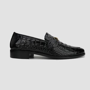 May include: A black, patent leather loafer with a crocodile skin texture. The shoe has a low heel and a gold-colored metal accent on the vamp. The shoe is designed for formal or business wear.