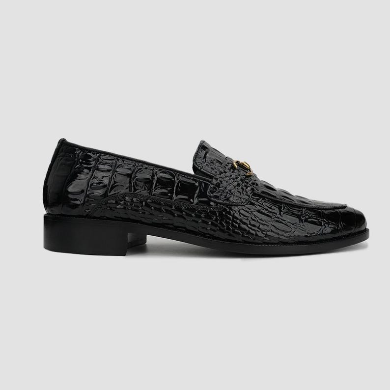 May include: A black, patent leather loafer with a crocodile skin texture. The shoe has a low heel and a gold-colored metal accent on the vamp. The shoe is designed for formal or business wear.