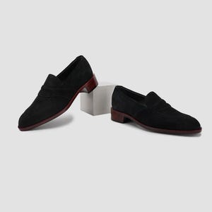 Puede incluir: Un par de mocasines negros de ante con suela y tac&oacute;n color burdeos. Los zapatos tienen un dise&ntilde;o cl&aacute;sico sin cordones con punta redondeada y una correa decorativa. Los tacones miden aproximadamente 2,5 cm de alto.