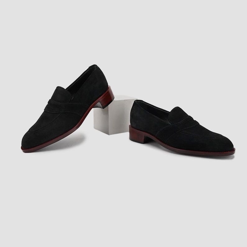 Puede incluir: Un par de mocasines negros de ante con suela y tac&oacute;n color burdeos. Los zapatos tienen un dise&ntilde;o cl&aacute;sico sin cordones con punta redondeada y una correa decorativa. Los tacones miden aproximadamente 2,5 cm de alto.