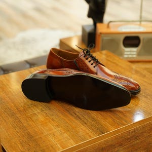 Puede incluir: Un par de zapatos de vestir de cuero marr&oacute;n con cordones negros, exhibidos sobre una superficie de madera. Los zapatos tienen un dise&ntilde;o cl&aacute;sico con perforaciones decorativas. Se ve la suela de un zapato, mostrando su color oscuro.