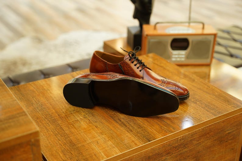 Puede incluir: Un par de zapatos de vestir de cuero marr&oacute;n con cordones negros, exhibidos sobre una superficie de madera. Los zapatos tienen un dise&ntilde;o cl&aacute;sico con perforaciones decorativas. Se ve la suela de un zapato, mostrando su color oscuro.