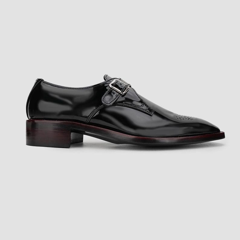 May include: A black leather dress shoe with a buckle closure. The shoe has a low heel and a pointed toe with decorative perforations. The sole has a dark red trim.