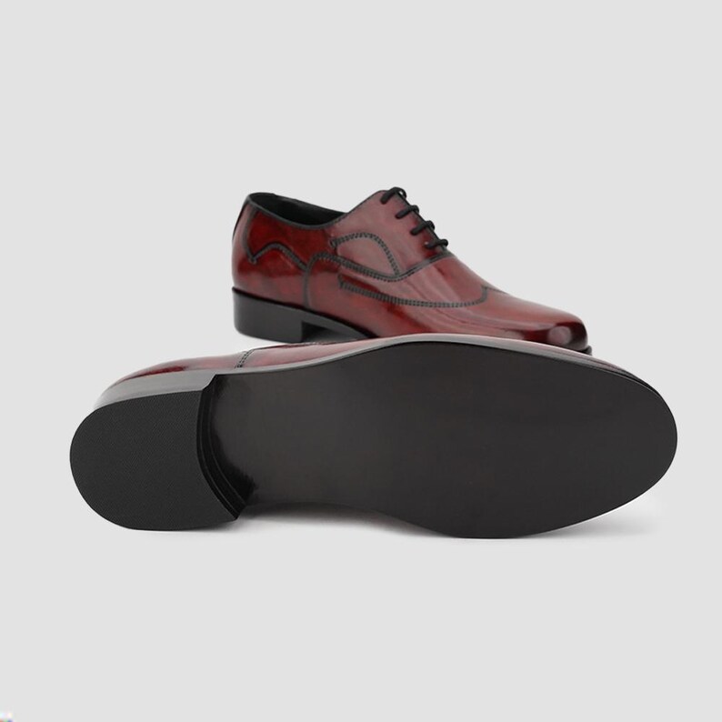 May include: A pair of burgundy leather dress shoes with black laces and stitching. One shoe is upright, and the other is positioned to show the sole. The shoes have a glossy finish and a classic wingtip design.