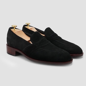 Puede incluir: Un par de mocasines negros de ante con suela y tac&oacute;n marr&oacute;n oscuro. Los zapatos tienen un dise&ntilde;o cl&aacute;sico con punta redondeada y una correa en la parte superior. Hormas de madera dentro de los zapatos.