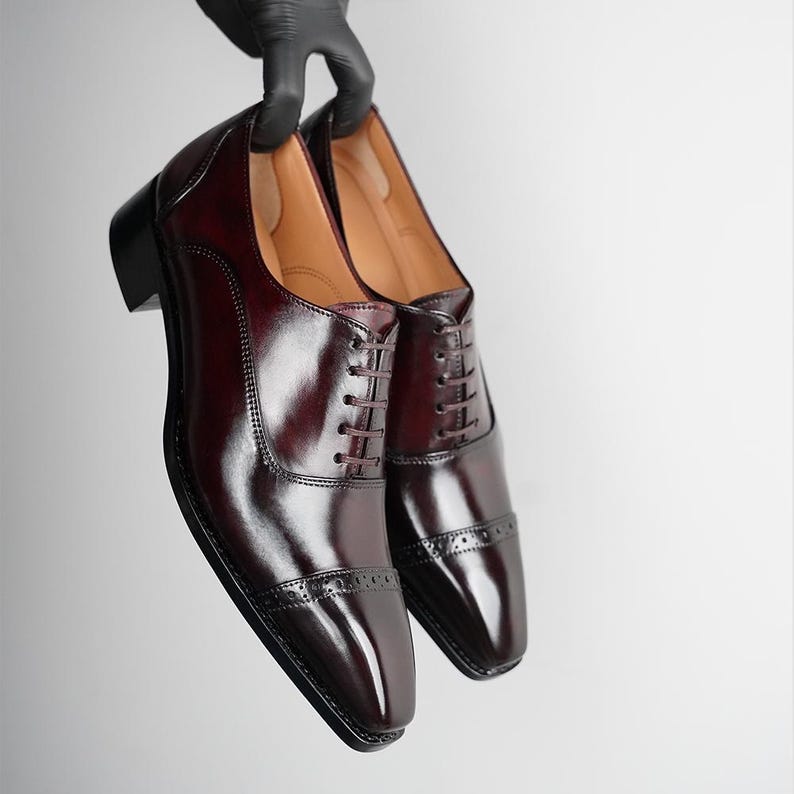 May include: A pair of polished oxblood leather oxford shoes featuring a cap toe with decorative brogue perforations, a lace-up front with thin dark laces, a stacked heel, and a smooth tan interior lining, presented in a sleek, formal design.
