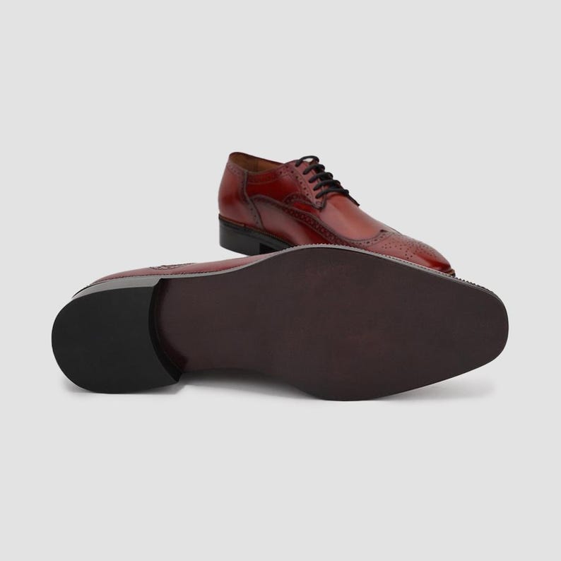Puede incluir: Zapatos de vestir de cuero burdeos con cordones negros. Los zapatos presentan un dise&ntilde;o de ala con perforaciones decorativas. Se ve la suela de un zapato, con color marr&oacute;n oscuro y tac&oacute;n.