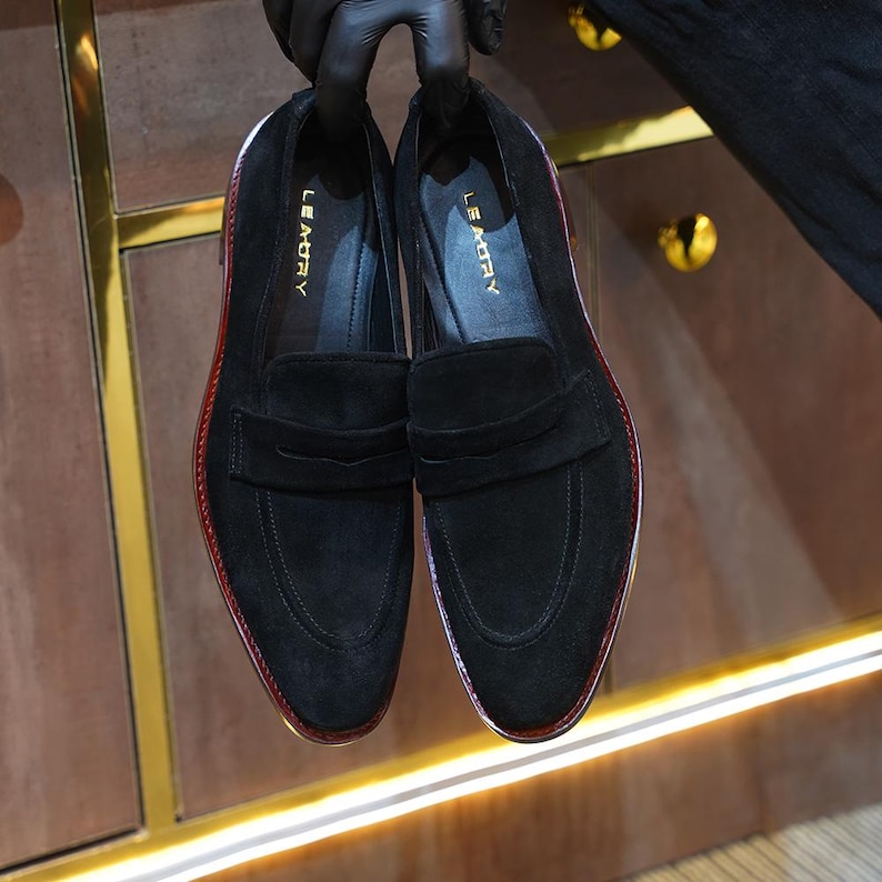 May include: A pair of black suede loafers with a penny strap design. The shoes have a red trim around the sole and the brand name "LE MORY" is visible on the insole. The shoes are held up by a person wearing black gloves.
