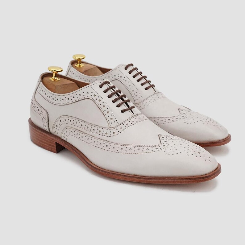 May include: A pair of white leather Oxford shoes with brown laces and a perforated design. The shoes have a wooden sole and heel, and are displayed with wooden shoe trees. The shoes are a classic style.