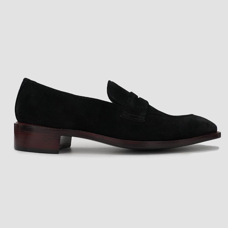 May include: Black suede loafer with a low, dark brown heel. The shoe features a classic penny loafer design with a rounded toe and a small strap detail across the top. The sole and heel are a contrasting dark brown color.