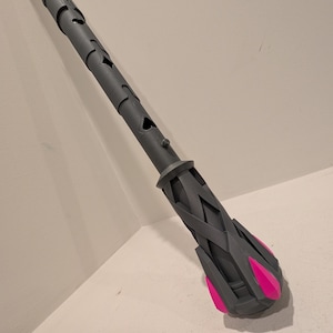 May include: A grey staff with a geometric design and pink accents. The staff has a series of cut-out shapes along its length and a woven pattern near the bottom. The end of the staff features a cluster of pink, petal-like shapes.