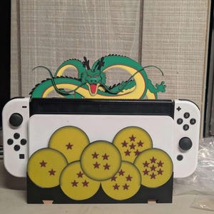 Może przedstawiać: Czarno-biała konsola Nintendo Switch z niestandardową stacją dokującą z zielono-żółtym wzorem smoka i żółtymi kulami z czerwonymi gwiazdami. Stacja dokująca została zaprojektowana tak, aby utrzymać konsolę w pozycji pionowej.