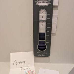 May include: A grey reading goal chart with the text "Goal: Read 10 Books" and "You can do it!". The chart has slots for tracking progress, with numbers 5, 6, 7, and 9 visible. A card reads "Great Job!" and a sticker says "Be Kind".