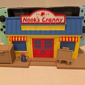 以下が含まれることがあります： 赤いと白のストライプのひさし、青い屋根、赤いドアを備えた、Animal CrossingのNook's Crannyのカラフルな3Dプリントモデル。Nintendo Switchがモデルの後ろに配置されています。茶色の箱、じょうろ、小さなイーゼルが含まれています。