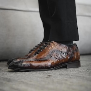 May include: Brown leather dress shoes with a textured, crocodile-like pattern. The shoes have a classic Oxford design with black laces and a dark brown sole. The toe cap is a darker shade of brown.
