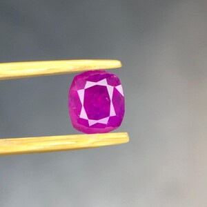 May include: A vibrant, cushion-cut magenta gemstone held by a pair of gold-toned metal tweezers. The stone features a faceted surface that reflects light, creating a geometric pattern across its deep pink, translucent body and polished edges.