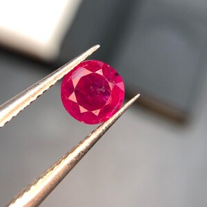 May include: A round, faceted, vibrant red gemstone held by metal tweezers. The stone features a brilliant cut with multiple triangular and kite-shaped facets that reflect light, creating a deep crimson colour with visible internal clarity characteristics.