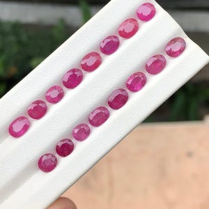 Könnte beinhalten: Eine Sammlung von fünfzehn oval geschliffenen, facettierten Rubinen, die in zwei parallelen Reihen auf einer weißen Präsentationskarte angeordnet sind. Jeder Edelstein zeigt einen leuchtenden, tiefrosa-roten Farbton.