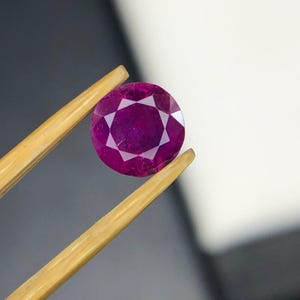 May include: A round, faceted, deep magenta-colored gemstone with a star-like pattern visible on its surface. The stone is held by a pair of light-colored wooden tweezers, showcasing its translucent quality and precise geometric cut.
