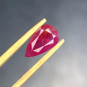 May include: A pear-shaped, faceted ruby gemstone with a deep red color and visible internal inclusions. The gemstone is held between two light-colored wooden tweezers, showcasing its translucent quality and geometric cut against a neutral background.