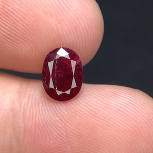 May include: An oval-cut, deep red ruby gemstone featuring a faceted surface with a distinct star-like pattern reflecting light. The stone displays a rich, saturated crimson hue with visible internal inclusions and sharp, geometric facets.