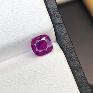Può includere: Una pietra preziosa taglio cuscino con una vibrante tonalità magenta-rosa e superfici sfaccettate che riflettono la luce. La pietra è esposta all'interno del rivestimento bianco testurizzato di un portagioie nero, mostrando il suo colore profondo.