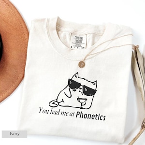 May include: Ivory-colored t-shirt with a black graphic of a cat wearing sunglasses and holding a drink. The text "You had me at Phonetics" is below the cat. A gold necklace and a brown hat are also in the image.