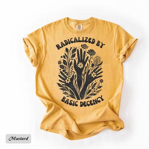 May include: Mustard-colored t-shirt with the text "RADICALIZED BY BASIC DECENCY" in a black, stylized font. The graphic features black hands and floral elements. The shirt is made of a soft material.