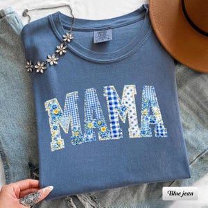 以下が含まれることがあります： 青色のComfort Colors Tシャツ。パッチワーク文字で「MAMA」と書かれています。文字は青と黄色の花柄とギンガムチェック柄です。花のアクセントが付いたシルバーのネックレスと茶色の帽子も写っています。