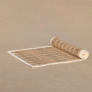 Puede incluir: Una esterilla de bamb&uacute; rectangular enrollada con un borde blanco. La esterilla est&aacute; parcialmente desenrollada, revelando un patr&oacute;n tejido de finas tiras de bamb&uacute; marr&oacute;n claro. La secci&oacute;n enrollada muestra el mismo material de bamb&uacute;.