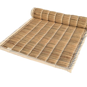 Puede incluir: Una esterilla rectangular enrollada, hecha de fibras naturales tejidas. La esterilla es principalmente beige con un borde beige claro y una secci&oacute;n enrollada en la parte superior. La textura tejida es visible, y la esterilla parece dise&ntilde;ada para la comodidad y la relajaci&oacute;n.