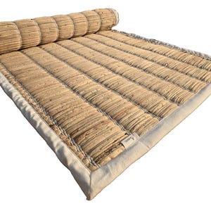 Puede incluir: Una esterilla rectangular enrollada, hecha de fibras naturales tejidas. La esterilla es de color beige claro con un borde blanco. La secci&oacute;n enrollada es cil&iacute;ndrica y cosida. Una peque&ntilde;a etiqueta es visible en la esterilla.