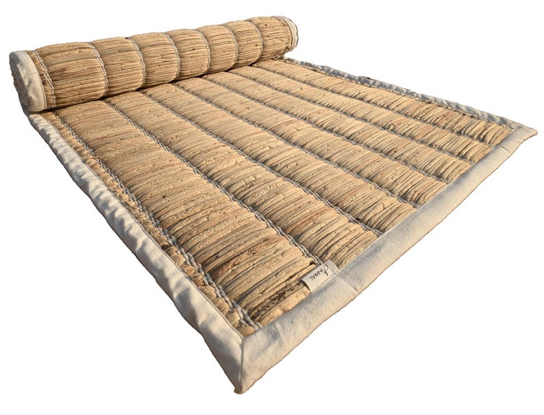 Puede incluir: Una esterilla rectangular enrollada, hecha de fibras naturales tejidas. La esterilla es de color beige claro con un borde blanco. La secci&oacute;n enrollada es cil&iacute;ndrica y cosida. Una peque&ntilde;a etiqueta es visible en la esterilla.
