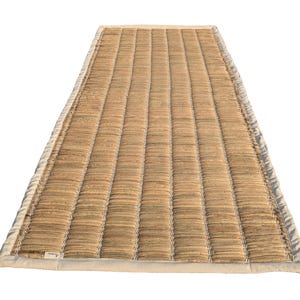 Puede incluir: Una estera rectangular tejida con una superficie texturizada de color marr&oacute;n claro. La estera tiene una serie de l&iacute;neas verticales paralelas y est&aacute; bordeada por una tela de color crema. La estera est&aacute; dise&ntilde;ada para su uso como revestimiento de suelo.