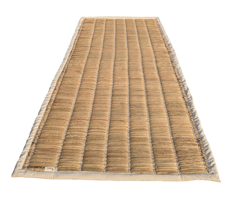 Puede incluir: Una estera rectangular tejida con una superficie texturizada de color marr&oacute;n claro. La estera tiene una serie de l&iacute;neas verticales paralelas y est&aacute; bordeada por una tela de color crema. La estera est&aacute; dise&ntilde;ada para su uso como revestimiento de suelo.
