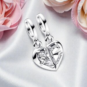 May include: Two silver heart-shaped pendants, each with a clear, sparkling gemstone, are displayed on a white satin surface. The pendants are split in half, suggesting a matching set. Pink and cream roses are in the background.