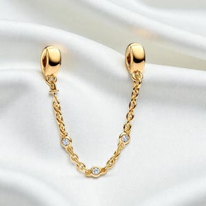 Może przedstawiać: Złoty łańcuszek zabezpieczający do zawieszek o kształcie litery U. Łańcuszek posiada małe, przezroczyste kamienie szlachetne i łączy się z dwoma złotymi końcówkami. Biżuteria jest prezentowana na białym, jedwabistym tle.